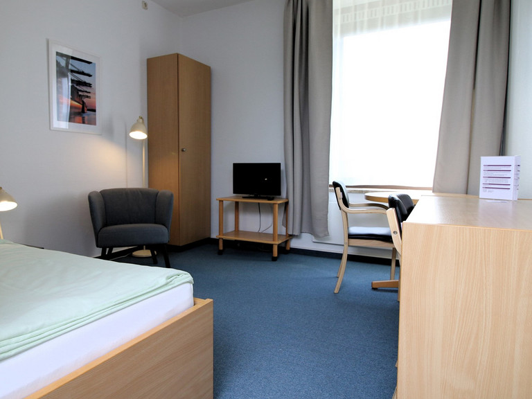Blick in ein helles Gästezimmer mit blauem Teppich, Bett, Tisch, Sessel und einem großen Fenster der Albertinen Akademie in Hamburg für die Unterkunft bei Tagungen, Fortbildungen, Weiterbildungen im Gesundheitswesen sowie zu touristischen Buchung. Blick in ein helles Gästezimmer mit blauem Teppich, Bett, Tisch, Sessel und einem großen Fenster der Albertinen Akademie in Hamburg für die Unterkunft bei Tagungen, Fortbildungen, Weiterbildungen im Gesundheitswesen sowie zu touristischen Buchung.